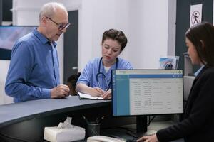 enfermeira olhando às médico relatório com idosos paciente enquanto assinatura documentos antes começar exame dentro hospital esperando sala. Senior homem tendo checar Visita com doutor, saúde Cuidado serviço foto