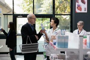 médico loja empregado avisando velho cliente protetor solar creme, falando com cliente dentro farmacêutico corredor. idosos comprador e jovem farmacêutico assistente discutindo protetor solar perto medicamentos estante foto