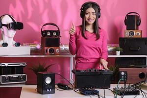 positivo músico em pé às dj mesa fazendo polegares acima gesto enquanto mistura eletrônico música e techno usando misturador console. ásia artista jogando estéreo sons com eletrônicos e microfone foto