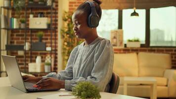 trabalhador autonomo trabalho às escrivaninha com música, desfrutando conectados trabalho tarefas enquanto ouvindo para canções em audio fone de ouvido. africano americano mulher teletrabalho às pôr do sol, escrevendo relatórios em bloco de anotações. foto