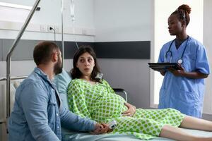 futuro pais discutindo com médico assistente dentro hospital ala, grávida mulher deitado dentro cama tendo contrações. africano americano enfermeira preparando paciente com gravidez para cirurgia foto