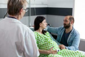 futuro pais discutindo trabalho processo com médico dentro hospital ala, paciente com gravidez deitado dentro cama preparando para médico cirurgia. grávida mulher segurando mãos em barriga ser reconfortante de homem foto