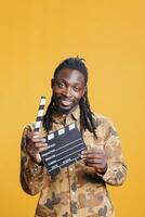 retrato do sorridente africano americano homem segurando filme claquete em pé dentro estúdio sobre amarelo fundo. jovem adulto dando audição para entretenimento vídeo. cinematográfico conceito foto