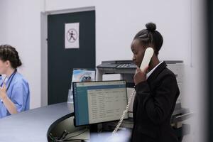 recepcionista falando às telefone fixo telefone com controlo remoto paciente discutindo médico compromisso detalhes enquanto em pé às hospital recepção contador. saúde Cuidado Apoio, suporte serviço e conceito foto