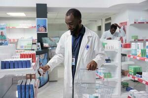 africano americano farmacêutico colocando Comida suplementos em farmácia prateleira, segurando compras cesta cheio do medicamentos. medicações varejo, farmacia loja mercadoria conceito foto