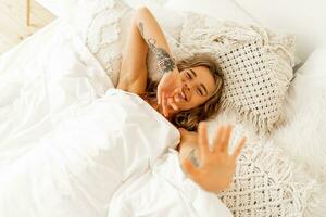 topo Visão retrato do feliz alegre mulher deitado dentro branco acolhedor cama despertar acima dentro a manhã, puxa mãos avançar. caloroso cores, boho interior. foto