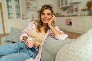 adorável mulher dentro casual roupas em repouso un sofá dentro acolhedor vivo sala. segurando copo do cappuccino. foto