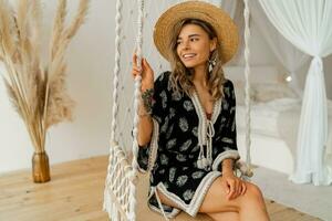 sorrindo jovem mulher dentro boho vestir posando dentro à moda quarto. palha chapéu, boho pena solta brincos . senhora sentado em suspensão balanço. foto