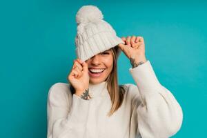 estúdio inverno retrato do sorridente loiro mulher dentro branco chapéu e grito suéter posando dentro estúdio sobre turquose fundo. frio estação acessórios e moda. foto