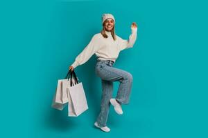 estúdio inverno retrato do sorridente loiro mulher dentro branco chapéu e grito suéter uma segurando compras bolsas , posando dentro estúdio sobre turquose fundo. cheio comprimento. foto