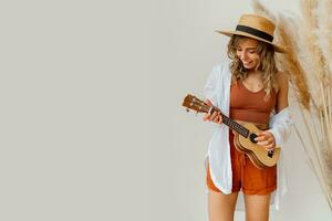 sorridente gracioso mulher dentro verão equipamento com Palha chapéu jogando ukulele guitare sobre branco fundo dentro estúdio com pampas Relva decoração. foto