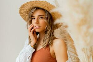 fechar acima retrato do atraente mulher dentro verão equipamento com Palha chapéu posando sobre branco fundo dentro estúdio com pampas Relva decoração. foto