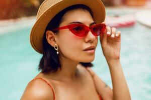 fechar acima retrato do à moda morena mulher dentro vermelho gato olhos oculos de sol relaxante dentro piscina durante tropical período de férias. foto