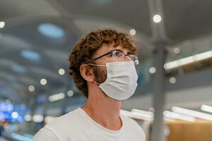 viajante homem vestindo face mascarar esperando para borda informação avião, em pé dentro saída terminal dentro aeroporto. foto