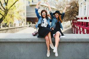 estilo de vida imagem do dois jovem bonita amigos meninas tendo Diversão ao ar livre em a rua. sentado em a ponte, conversação, rindo, levantar mãos acima. positivo humor. amizade. foto