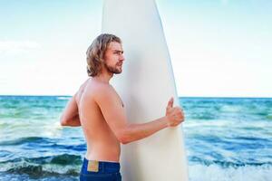 jovem atraente surfista Esportes homem com grandes cabelo vestindo jeans calção e olhando às a horizonte enquanto carregando dele surfar borda durante uma ensolarado dia contra a intenso azul céu. foto