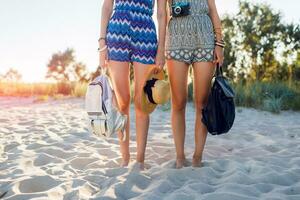 ao ar livre tiro do alegre jovem fêmea amigos caminhando juntos em uma de praia às pôr do sol. dois atraente mulheres desfrutando uma feriado em a mar costa. vestindo na moda verão roupa, mochilas , oculos escuros. foto