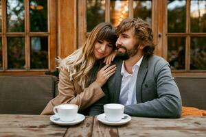 romântico pensativo mulher com grandes ondulado cabelos abraçando dela marido com barba. elegante casal sentado dentro cafeteria com quente cappuccino. foto