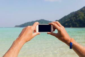 homem mãos segurando branco Móvel telefone com Preto tela. surpreendente azul mar e céu tropical ilha fundo. tailândia. foto