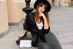 confiante à moda morena mulher posando dentro europeu cidade, caminhando em a rua. vestindo casual roupa, Preto chapéu e blusa. outono moda olhar. foto