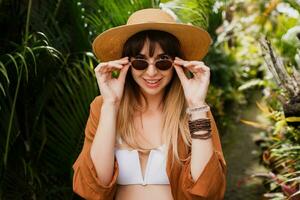 fechar acima verão elegante retrato do morena mulher dentro Palha chapéu posando em tropical Palma folhas fundo dentro bali. vestindo à moda boêmio acessórios. foto