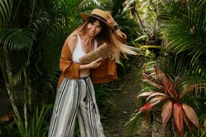 moda retrato do morena mulher dentro Palha chapéu posando em tropical Palma folhas fundo dentro bali. vestindo à moda boêmio acessórios. foto