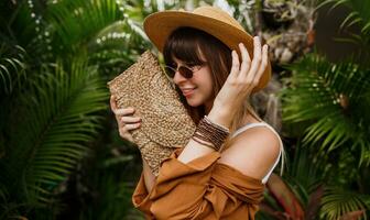 fechar acima verão elegante retrato do morena mulher dentro Palha chapéu posando em tropical Palma folhas fundo dentro bali. vestindo à moda boêmio acessórios. foto