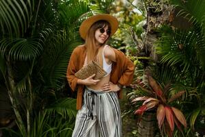 moda imagem do sexy gracioso mulher dentro Palha chapéu posando em tropical Palma folhas fundo dentro bali. vestindo à moda boêmio acessórios. foto