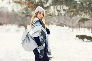 inverno estilo de vida retrato do jovem Loiras senhora dentro caloroso lã fofo cachecol e chapéu com mochila caminhando dentro Maravilhoso inverno parque. foto