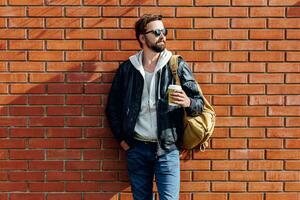 ao ar livre outono ou inverno retrato do bonito hipster homem com barba , cinzento moletom com capuz e couro Jaqueta com saco pacote segurando copo do quente café. tijolo urbano parede fundo. foto