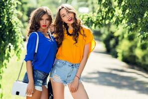 dois lindo meninas caminhando dentro a verão parque fim conversando. amigos vestindo à moda camisa e jeans calção , desfrutando dia fora e ter Diversão. foto