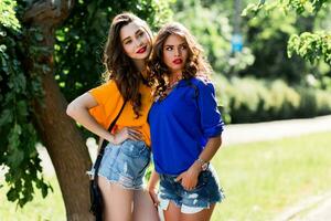 dois lindo meninas caminhando dentro a verão parque fim conversando. amigos vestindo à moda camisa e jeans calção , desfrutando dia fora e ter Diversão. foto