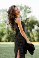 verão moda retrato do r elegante mulher com perfeito ondulado cabelo dentro à moda elegante Preto chapéu e brilhante faço acima posando dentro a parque. rua estilo. foto