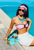 estilo de vida retrato do bronzeada lindo mulher dentro Rosa bikini e oculos de sol sentado perto natação piscina com fresco coquetel. surpreendente sensual menina com perfeito fino esporte corpo posando ar livre. foto