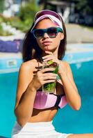 estilo de vida retrato do bronzeada lindo mulher dentro Rosa bikini e oculos de sol sentado perto natação piscina com fresco coquetel. surpreendente sensual menina com perfeito fino esporte corpo posando ar livre. foto