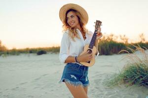 bem torneado gengibre senhora jogando ukulele guitarra dentro Sol luz em a de praia . vestindo Palha chapéu e na moda jeans shorts. caloroso pôr do sol cores. foto