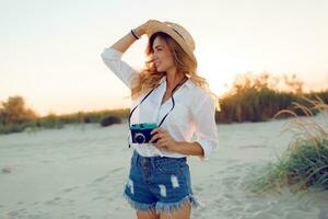 viajando e feriados conceito. feliz fêmea caminhando em tarde de praia e segurando retro Câmera. vestindo Palha chapéu e sexy jeans shorts. caloroso pôr do sol cores . foto