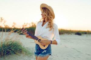 bem torneado gengibre senhora jogando ukulele guitarra dentro Sol luz em a de praia . vestindo Palha chapéu e na moda jeans shorts. caloroso pôr do sol cores. foto