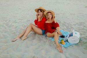 dois bonita mulheres dentro vermelho verão equipamento abd Palha chapéus desfrutando piquenique em a de praia. verão humor. foto