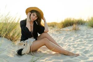 alegre morena mulher dentro à moda verão boho equipamento posando em a de praia. pôr do sol cores. Palha chapéu. foto