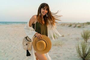 verão foto do feliz jovem mulher dentro à moda boho equipamento segurando Palha saco e chapéu posando em tropical de praia. ventoso cabelos.