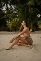 lindo bronzeada mulher sentado e banhos de sol às a de praia. elegante fino garota. ao ar livre foto. verão vibrações. à moda roupa de banho. foto