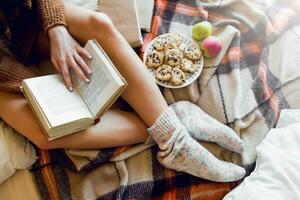 suave acolhedor foto do fino bronzeado mulher dentro caloroso suéter em a cama com boock dentro mãos, topo Visão ponto. menina sentado em xadrez xadrez perto velho livros.