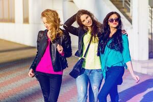 três melhor amigos ter Diversão em ensolarado outono dia urbano rua fundo . grupo do alunos andando, vestindo cor outono suéter e couro jaqueta. brilhante cores. foto