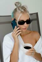 fechar acima retrato do sorridente loiro mulher dentro à moda oculos de sol e branco roupão bebendo copo do café dentro cedo manhã. em repouso dentro luxo hotel. foto