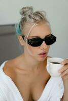 fechar acima retrato do prazer loiro mulher dentro à moda oculos de sol e branco roupão bebendo copo do café dentro cedo manhã. foto