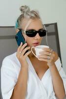 fechar acima retrato do sorridente loiro mulher dentro à moda oculos de sol e branco roupão bebendo copo do café dentro cedo manhã. em repouso dentro luxo hotel. foto