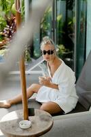 verão foto do prazer loiro mulher dentro à moda oculos de sol e branco roupão bebendo copo do café dentro cedo manhã.