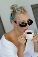 fechar acima retrato do prazer loiro mulher dentro à moda oculos de sol e branco roupão bebendo copo do café dentro cedo manhã. foto