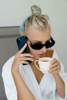 verão foto do alegre loiro mulher dentro à moda oculos de sol e branco roupão bebendo copo do café enquanto arrepiante dentro luxo hotel. falando de Móvel telefone.